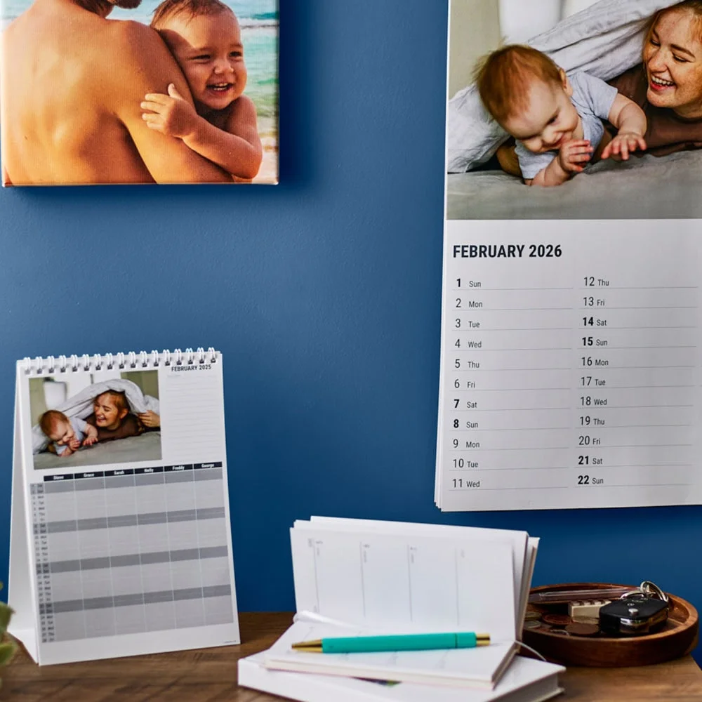 A desk setup with a blue wall featuring a photo calendar open to February 2026, a framed timeline print, and a photo canvas. On the desk are a small spiral calendar with a photo, a notebook with a pen, and a potted plant. The scene includes warm wooden tones and decorative greenery.